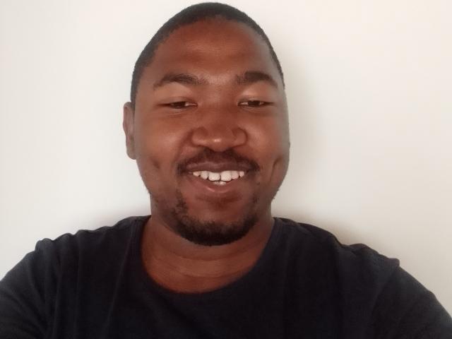 BigDickCya261 has a dark brown face, short black hair, and a small goatee. He's smiling with white teeth, wearing a black shirt. His eyes are half-closed.