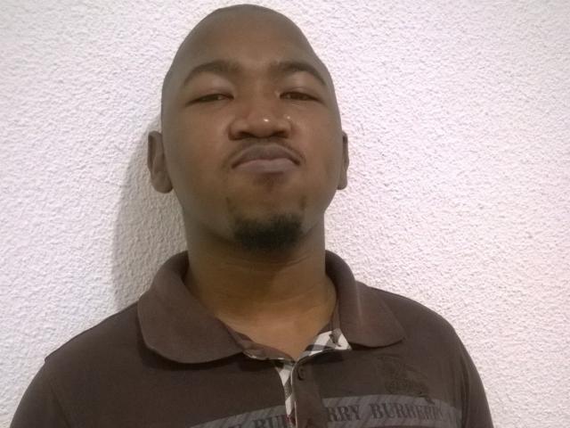 BigDickCya261, bald, medium brown skin, slight goatee, black polo shirt, looking straight at camera, subtle smirk, textured white wall in background.
