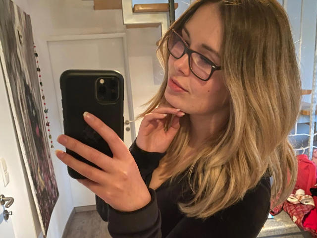 Shyla, mit blonden Haaren, Brille und einem schwarzen T-Shirt, macht ein Selfie vor einem Spiegel mit ihrem Handy, zeigt ein leichtes Lächeln und legt ihr Kinn in ihre Hand..