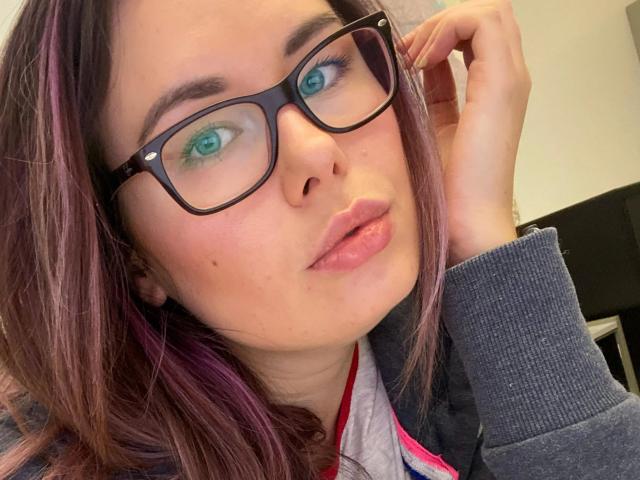 Shyla, sinnlich mit lila gefärbten Haaren, einer schwarzen Brille und sinnlichen, rosafarbenen Lippen, ihre grünen Augen blicken aufwärts, die Hand in der Nähe ihres Gesichts, trägt eine graue Jacke mit roten und pinkfarbenen Akzenten..
