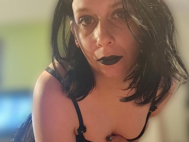 WorshipKate with black lipstick and black hair, wearing a black bra, leaning forward, showing her tits, nose ring, and dark makeup, looking into the camera.