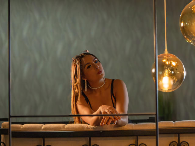 TiffanyMartins with long blonde dreads, wearing a black tank top, pearl necklace, and sunglasses on her head, leaning on a white cushioned bar, with gold hanging lights.