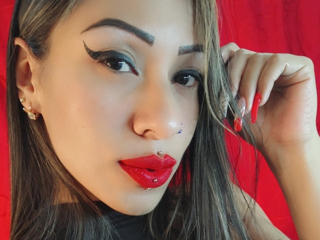 A LunaCandy tem batom vermelho, um piercing no nariz e um piercing no lábio. Ela está usando delineador preto com pontas para cima, unhas vermelhas e tem um fundo vermelho. O cabelo castanho dela está de um lado..