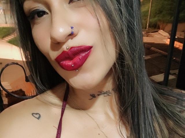 LunaCandy has long black hair, red lipstick, a nose ring, and a lip ring. She has tattoos on her neck and chest, with a heart tattoo on her shoulder.