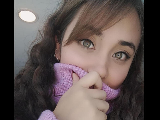 ArtemisNati with curly brown hair, green eyes, and a purple knitted turtleneck, holding it to her nose with her finger, showing her cute face and long eyelashes.