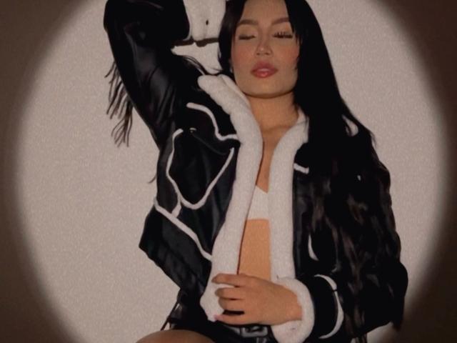 AshlyFerria with long black hair, wearing a black and white jacket, has her hand on her chest, exposing her tan-colored tits, with her eyes closed and lips slightly parted.