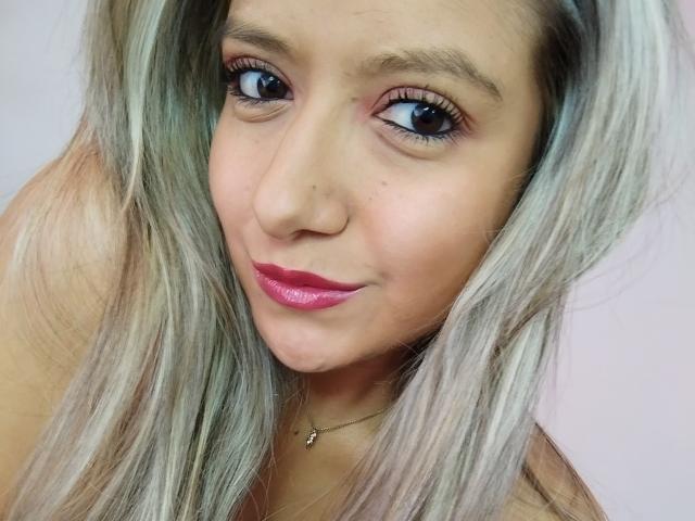 ValeriaHunter69 has light blonde hair, pink lipstick, and brown eyes, with a small nose and subtle makeup, wearing a necklace with a tiny cross, smiling slightly.