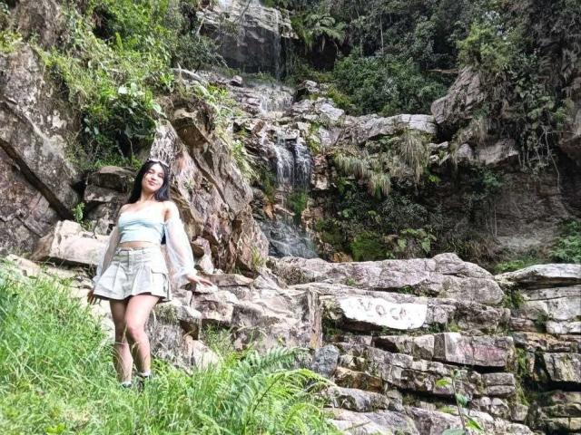 ValeriiNaughty has dark hair, light skin, small tits, wearing a light blue off-shoulder top and green skirt, standing by rocky waterfall, green grass in foreground.