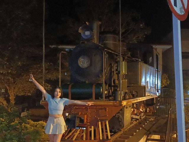 ValeriiNaughty with arms out, wearing a blue crop top and white skirt, standing next to an old steam locomotive at night, arms raised, lit by streetlight.
