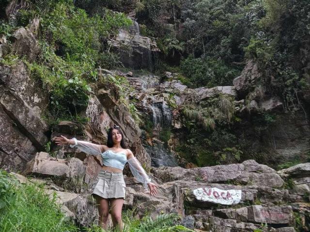 ValeriiNaughty stands with arms out, wearing a blue off-shoulder top and gray shorts, in a rocky, green forested area with a small waterfall. Her tits are visible.