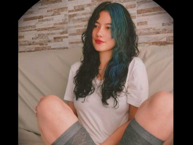 LauryLu with curly black and blue hair, red lipstick, white tee, and grey socks, sitting with knees up, smiling slightly. Her ass is partially visible.