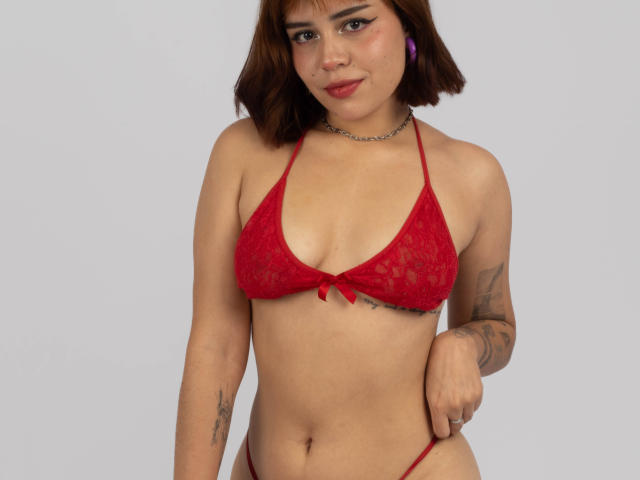 Anni Evans mit rotem Spitzen-Bikini, kleinen Brüsten, süßem Gesicht, braunen Bob-Frisur, Tattoos, lila Ohrringen, einer Kette um den Hals, einem leichten Lächeln, hält den Bikini-Unterteil, helle Haut, Studioaufnahme..
