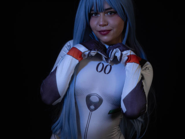 Anni Evans mit langen, blauen Haaren, trägt ein eng anliegendes, weißes und braunes Cosplay-Kostüm, ihre Hände sind in der Nähe ihres Gesichts, sie lächelt leicht, blaue Augen, sichtbare Brüste, dunkler Hintergrund..