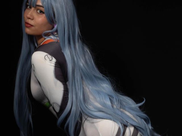 Anni Evans mit langen, blauen Haaren, in einem weißen und schwarzen Cosplay-Kostüm, leicht nach vorne gebeugt, zurückblickend, mit kleinen Brüsten und einem süßen Gesicht, vor schwarzem Hintergrund..