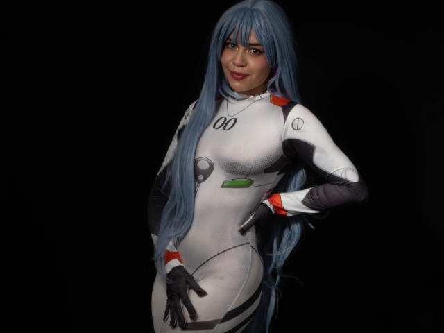 Anni Evans mit langen, blauen Haaren, trägt einen engen, weißen und grauen, futuristischen Body und steht vor einem schwarzen Hintergrund. Sie hat eine Hand in die Hüfte gestemmt, lächelt und präsentiert ihre kurvigen Hüften und ihre kleinen Brüste..