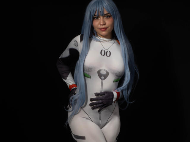 Anni Evans mit langen, blauen Haaren, in einem schwarz-weißen Cosplay-Kostüm, das ihre Brüste in einem engen Anzug zeigt, mit schwarzen Handschuhen, stehend mit den Händen in den Hüften, lächelnd, dunkler Hintergrund..