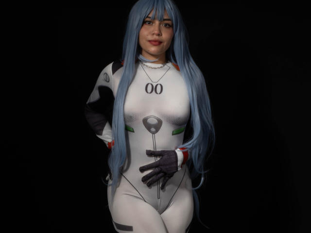 Anni Evans mit langen, blauen Haaren, trägt einen engen, weißen und grauen Cosplay-Anzug, ihre Hand liegt auf ihrem Intimbereich, sie trägt schwarze Handschuhe und steht vor einem dunklen Hintergrund, lächelnd..