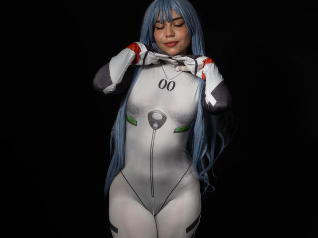 Anni Evans mit blauen Haaren in einem engen, weißen und grauen Cosplay-Body, wobei sie ihre Brüste mit den Händen bedeckt, steht vor einem schwarzen Hintergrund und lächelt mit geschlossenen Augen..