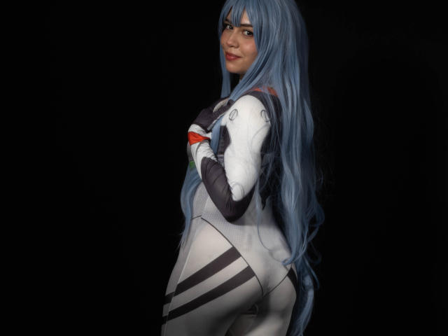 Anni Evans mit langen, blauen Haaren, trägt ein eng anliegendes Cosplay-Kostüm in Weiß und Schwarz, das ihren Hintern zeigt, steht seitlich und blickt mit einem leichten Lächeln zurück..