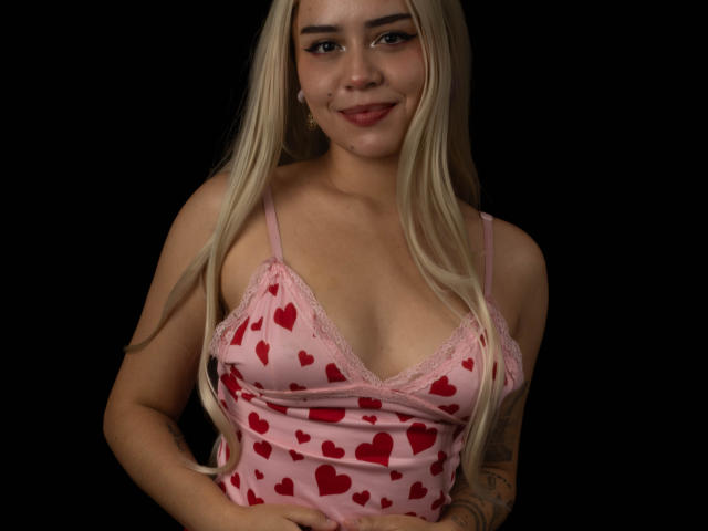 Anni Evans mit langen, blonden Haaren, trägt eine rosa Tanktop mit Herzmuster, lächelt, zeigt ihre Brüste, hat ein Tattoo am rechten Arm, vor einem schwarzen Hintergrund..