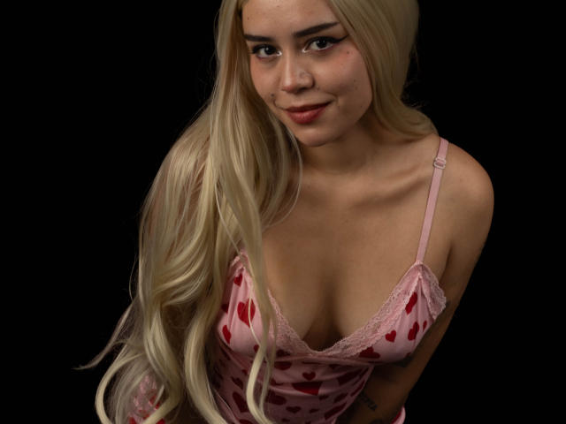 Anni Evans mit langen blonden Haaren, pinkfarbener Spitze mit Herzmuster und Spitzenbesatz, die ihre Brüste zeigt und sanft in die Kamera lächelt. Sie hat helle Haut und braune Augen..