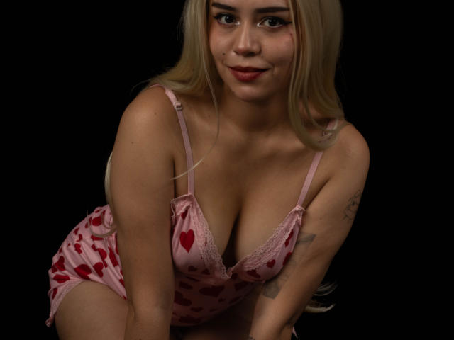 Anni Evans mit blonden Haaren, die rosige, herzmusterige Unterwäsche trägt, zeigt ihre Brüste, lehnt sich vor, lächelt, hat ein Tattoo am linken Arm, vor schwarzem Hintergrund..
