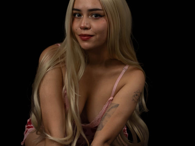 Anni Evans mit langen blonden Haaren, einem pinken Spitzen-BH, einem Tattoo am rechten Arm, sitzend, die Kamera anblickend, mit einem leichten Lächeln, vor schwarzem Hintergrund, bei weicher Beleuchtung, wobei ihre Brüste und ihr süßes Gesicht zu sehen sind..