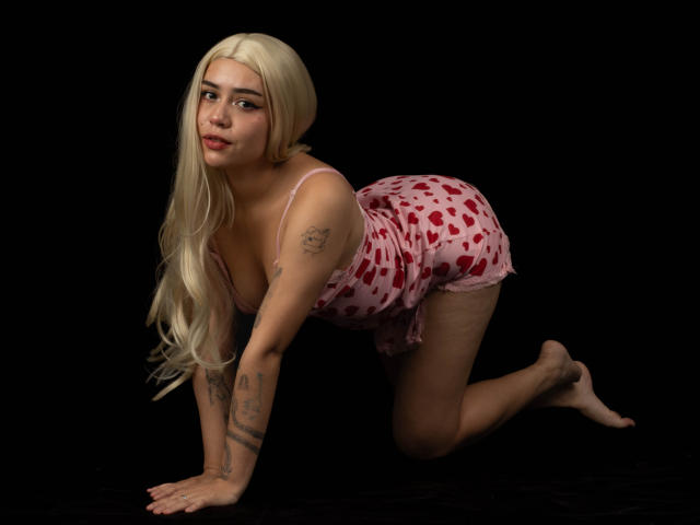 Anni Evans befindet sich auf allen Vieren, trägt ein pinkes Nachthemd mit Herzmuster, hat langes blondes Haar, Tattoos an den Armen und einen schmollenden Gesichtsausdruck, vor einem schwarzen Hintergrund..