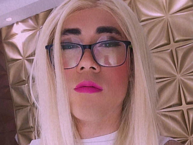 ElizabethQuenX with blonde wig, black glasses, pink lipstick, and white shirt, looking slightly pouty, has a clean, fair-skinned face, and is centered in the image.