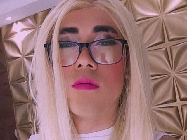 ElizabethQuenX with blonde hair, black glasses, pink lipstick, and a serious expression, showing her nose and slightly blushed face. She's wearing a white top, looking straight at camera.