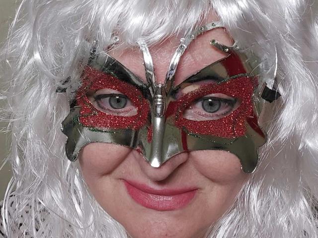MadyAllav has silver metallic and red glittery mask, white wavy wig, and pink lipstick, smiling with blue eyes. Her ass is not visible, just her face.