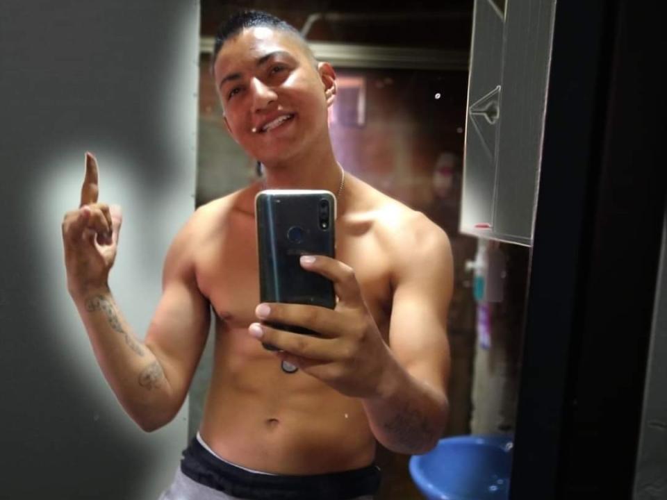 Angulus, torse nu avec une musculature tonique, prend un selfie devant un miroir. Il sourit, pointe du doigt avec son index droit, et a des tatouages sur son bras gauche..