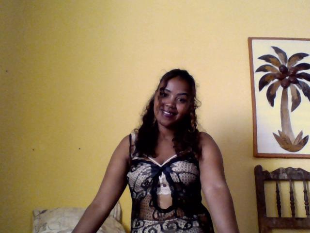 Nairahmy has dark curly hair, brown skin, and is smiling while wearing a black lace lingerie top that reveals her small boobs and a cut-out below her chest.
