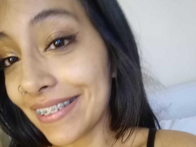 NoremCute tiene la piel morena, ojos marrón oscuro, cabello negro largo, un piercing en la nariz y está sonriendo, mostrando brackets metálicos en sus dientes. Lleva una blusa negra..
