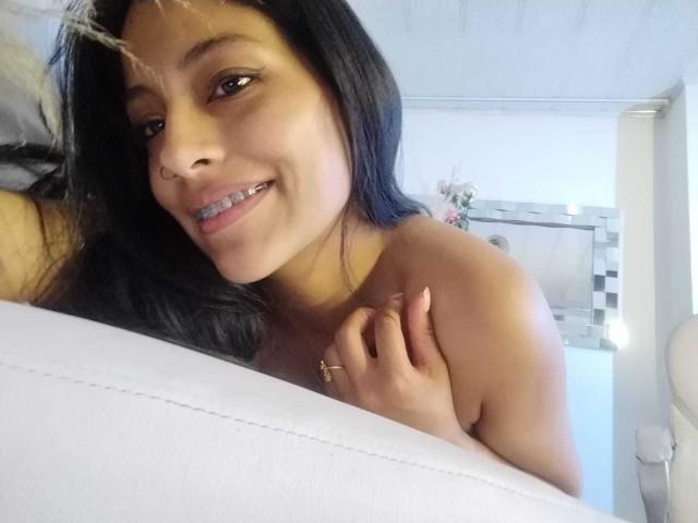 NoremCute, sonriendo con cabello negro, piercing en la nariz y diastema, sosteniendo un almohadón blanco, sin ropa en la parte superior, mostrando los hombros desnudos, usando un anillo, con una cara linda y una sonrisa bonita..