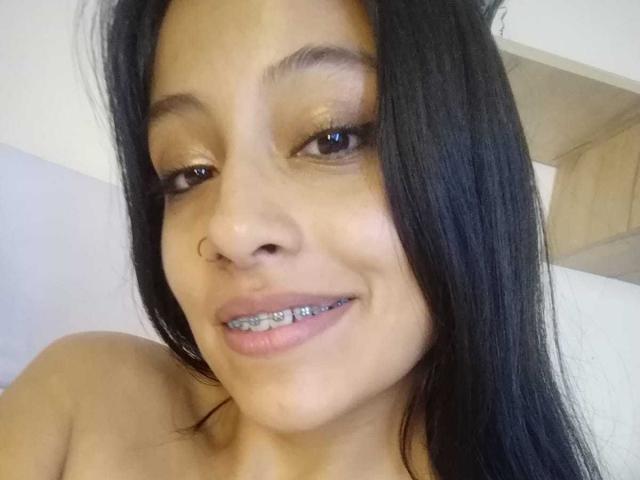 NoremCute has tan skin, black hair, and a nose ring. She's smiling with metal braces on her teeth. Her eyes are brown with shiny gold eyeshadow.