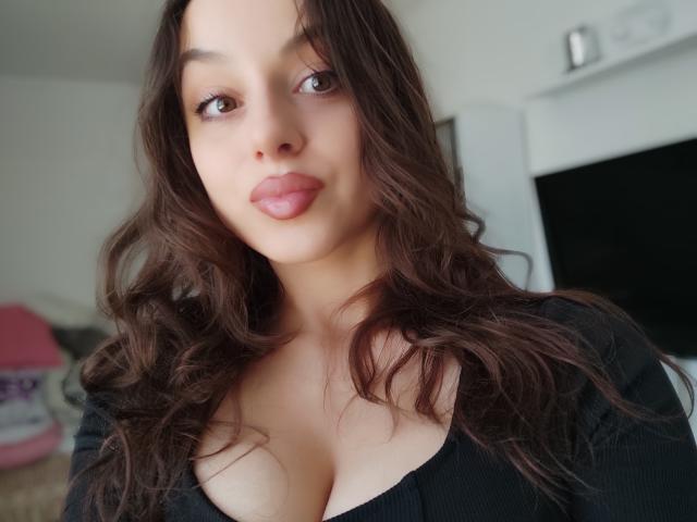 CourneyLust has wavy brown hair, brown eyes, and is wearing a black top with cleavage, showing her boobs. She has pink lips and a soft, sexy expression.