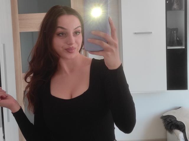 CourtneyLust with medium tits, wearing a black long-sleeve top, taking a selfie in a mirror, has long brown hair, a cute smile, and is holding a phone.