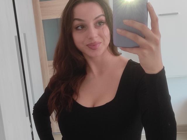 Selfie of CourtneyLust with wavy brown hair, black long-sleeve top, showing her tits in a mirror, holding a phone with a flash, slightly puckered lips, smiling.