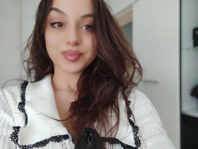 CourtneyLust with wavy brown hair, subtle makeup, and pink lips, wearing a white, black-trimmed shirt, looking directly at the camera with a gentle smile.