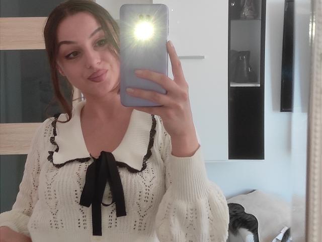 CourtneyLust with pouty lips, brown hair in a ponytail, white lace-up shirt, black ribbon, holding a phone with a bright flash, taking a mirror selfie. Boobs visible through shirt.