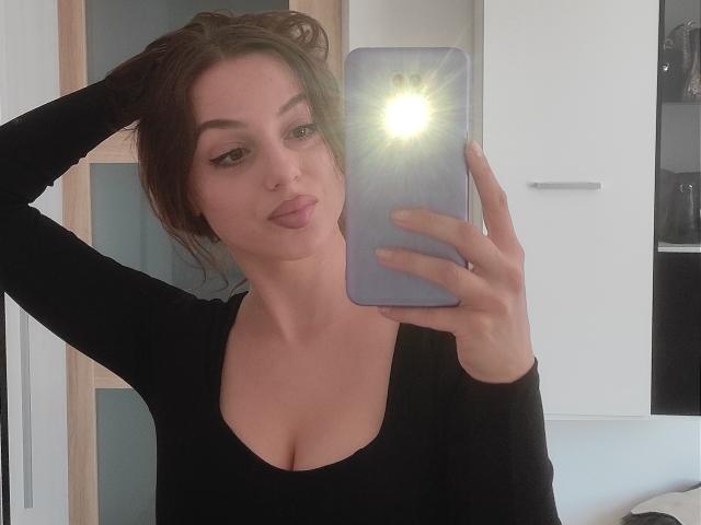 CourtneyLust with her hand in her brown hair, black top, and cleavage, holding a phone with a bright flash, looking at the camera with pouty lips.