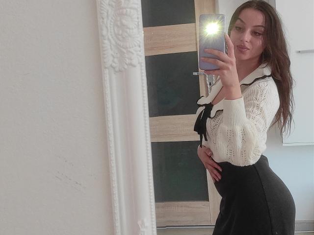 CourtneyLust with long brown hair, wearing a white lace blouse and black skirt, holding a phone in a mirror selfie, showing her tits and ass, in a white room.