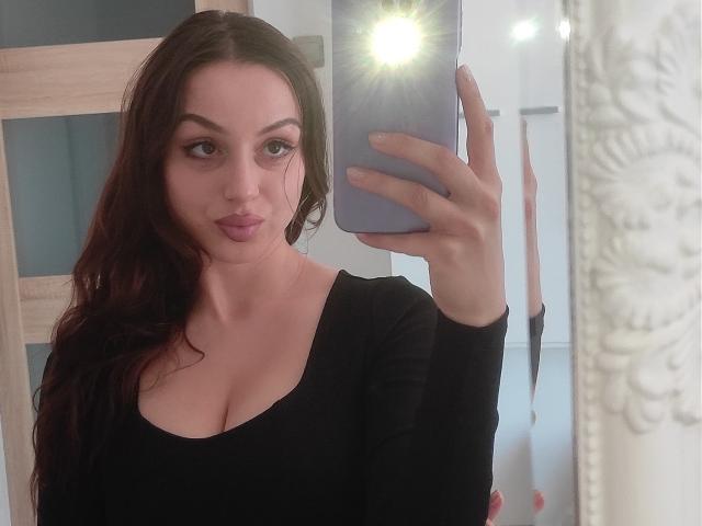 CourtneyLust with wavy brown hair, pouty lips, and big tits in a black top, taking a mirror selfie with a blue phone, reflection showing a bright light.