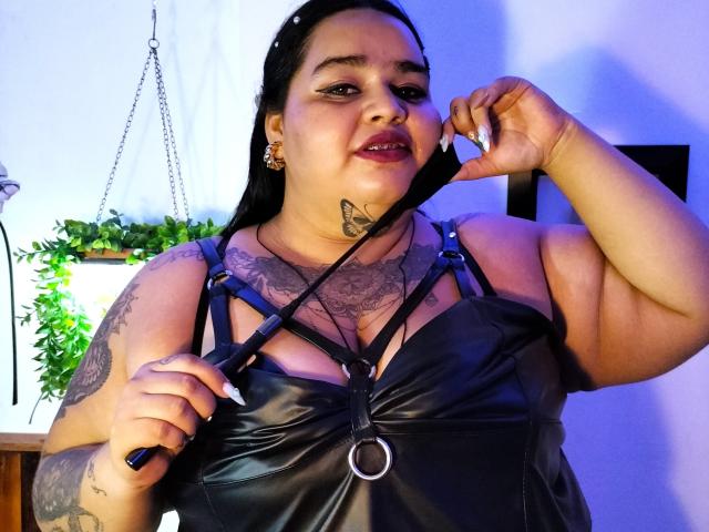 VeronikChubby with black leather dress, holding a black strap, showing tattoos, butterfly neck tattoo, and smiling, dark hair, blue eyes, purple lipstick, in a room with purple lighting.