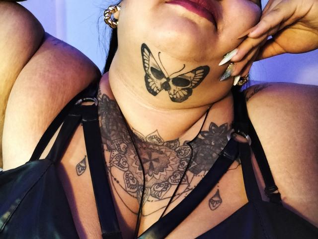 VeronikChubby's face, showing a butterfly tattoo on her neck and intricate chest tattoos, wearing black leather straps, with her hand near her mouth and long silver nails.