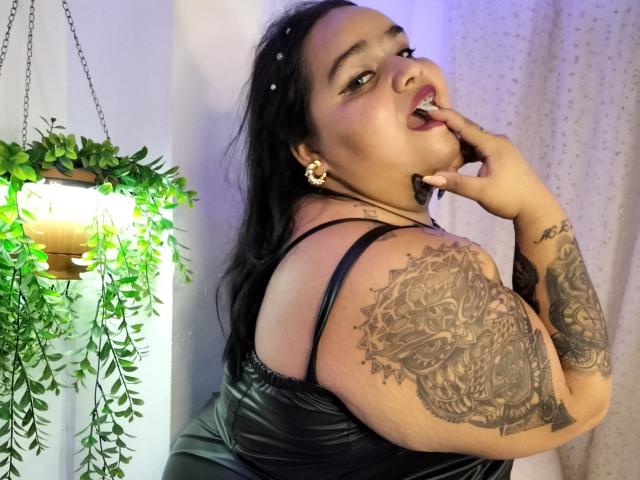 VeronikChubby with dark hair, tattoos on her arm, black leather dress, gold earrings, finger in mouth, green plant hanging left, white curtain right, purple lighting.