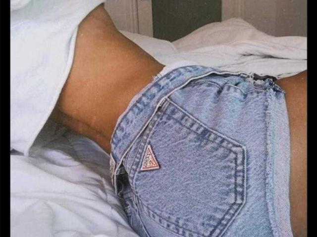 I glutei di UmaRany, in pantaloncini di jeans blu sbiaditi, mostrano una tasca Levi's a forma di triangolo. Camicia bianca tirata su, che rivela una sezione centrale sottile e abbronzata. Letto con lenzuola bianche sullo sfondo..