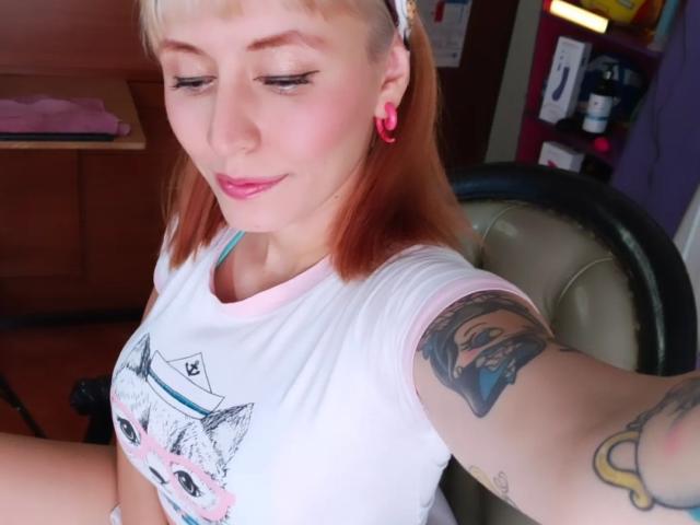 SaraleePoison with pink ear gauges, white shirt, and pink lips, showing a tattoo on her right arm, blonde hair, and small tits, sitting in an office chair.