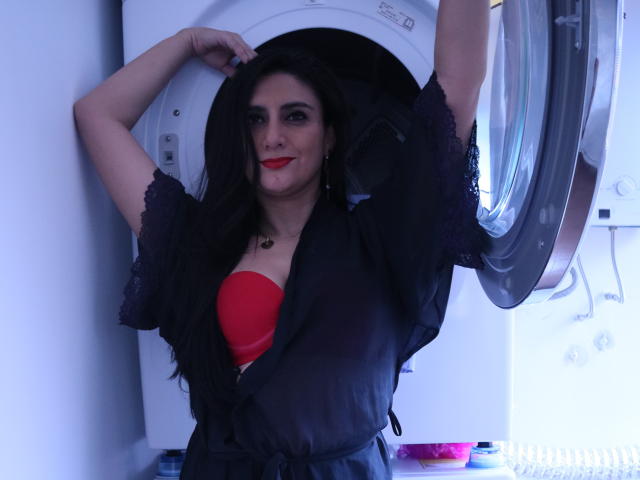 JuliaRobinson with black lace robe, red bra, and long black hair, standing in front of an open washing machine, arms raised, smiling with red lipstick.